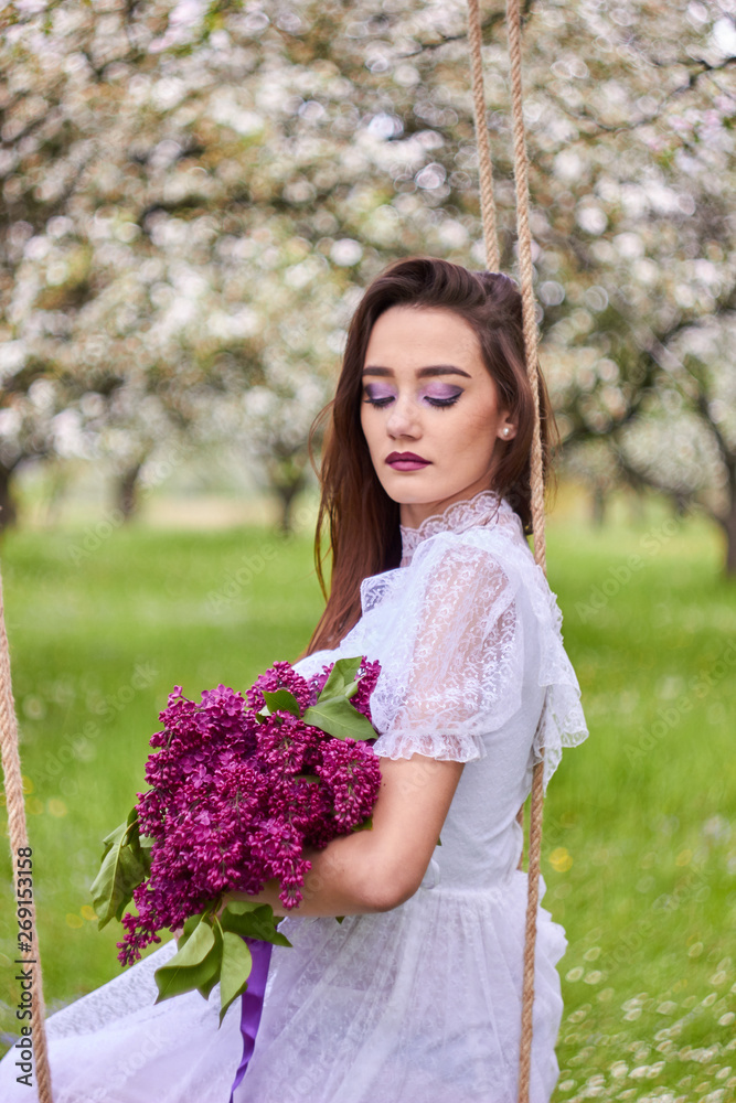 A young beautiful girl with a bouquet of lilac sits on a makeshift swing in an apple orchard in spring. White lace dress on the perfect figure of the model. Photo session in the open. A young beautiful girl with a bouquet of lilac sits on a makeshift swing in an apple orchard in spring. White lace dress on the perfect figure of the model. Photo session in the open.
