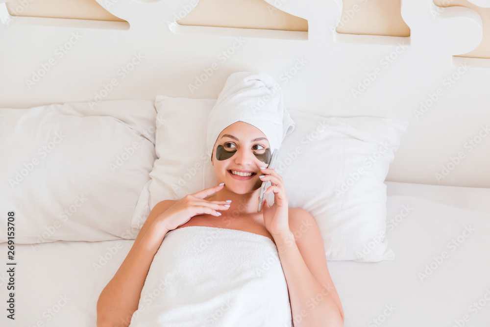 Beautiful smiling young woman with under eye patches and talking mobile phone in bathrobe lying in bed. Happy girl taking care of herself. Beauty skincare and wellness morning concept Beautiful smiling young woman with under eye patches and talking mobile phone in bathrobe lying in bed. Happy girl taking care of herself. Beauty skincare and wellness morning concept