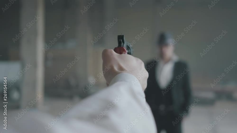 The hand of an unrecognizable man aiming a gun, blurred figure of a confident man in the suit and hat in front of him. The guy is going to kill his enemy. Mafia disassembly