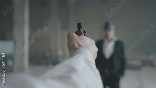 The hand of an unrecognizable man aiming a gun, blurred figure of a confident man in the suit and hat in front of him. The guy is going to kill his enemy. Mafia disassembly photo