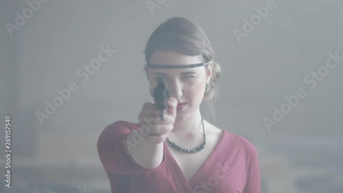 Confident attractive woman in stylish red dress is aiming a pistol at the camera in an abandoned dusty building, then lowers the gun. Dangerous lady. Retro, vintage style