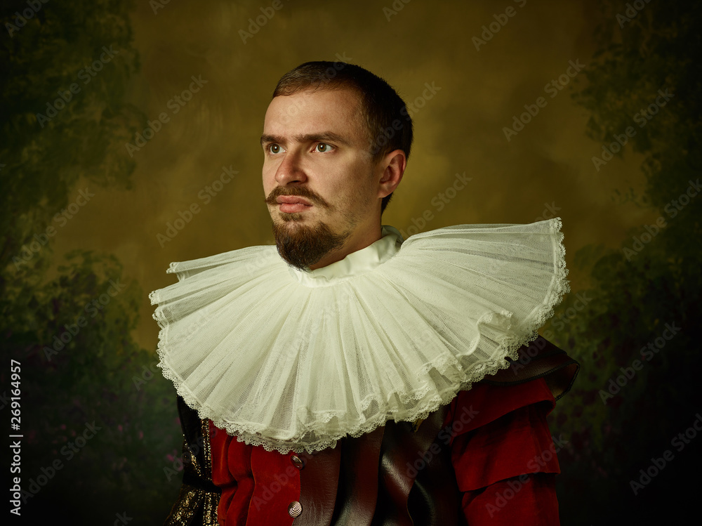 Young man as a medieval knight on dark studio background. Portrait in low  key of male model in retro costume. Looks serious. Human emotions,  comparison of eras and facial expressions concept. Stock