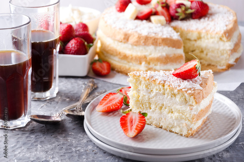 Closeup of a slice of sponge cake on the plate, glasses of coffee, bowl of strawberries, whole sponge cake on the background