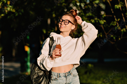 Happy student woman wearing white pullover and jeans with backpack holding a bottle with juice and walking in the park 