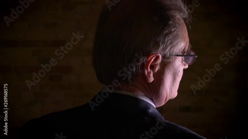 Close-up rear silhouette of senior man in costume watches dramatically standing in complete darkness.