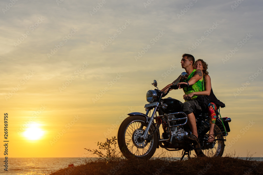 Romantic picture with a couple of beautiful stylish bikers at sunset. Handsome guy with tatoo and young sexy woman enjoy themselves in motorbike trip.