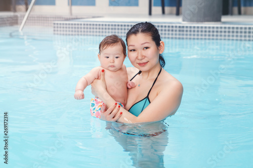 Mixed race asian mother holding her newborn baby in swimming pool standing in water and looking in camera. Mom with little child. Healthy active lifestyle. Family activity and bonding © anoushkatoronto