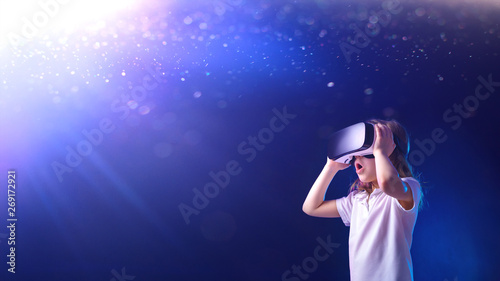 Surprised female kid using digital glasses of virtual reality on shining blue background