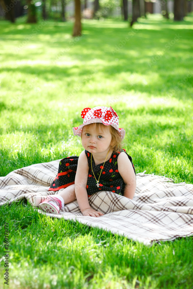 Happy little blonde girl-child in cherry pattern dress play in summer park under the trees shade of a beautiful green grass. Childhood. Concept of a happy family. Universal Children's Day Happy little blonde girl-child in cherry pattern dress play in summer park under the trees shade of a beautiful green grass. Childhood. Concept of a happy family. Universal Children's Day