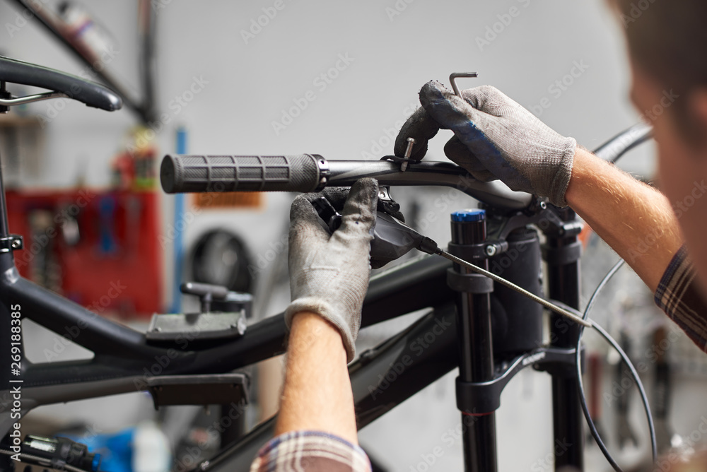Cropped shot of male worker working in bicycle repair shop, professional mechanic repairing mountain bike using special instrument, wearing protective gloves Cropped shot of male worker working in bicycle repair shop, professional mechanic repairing mountain bike using special instrument, wearing protective gloves