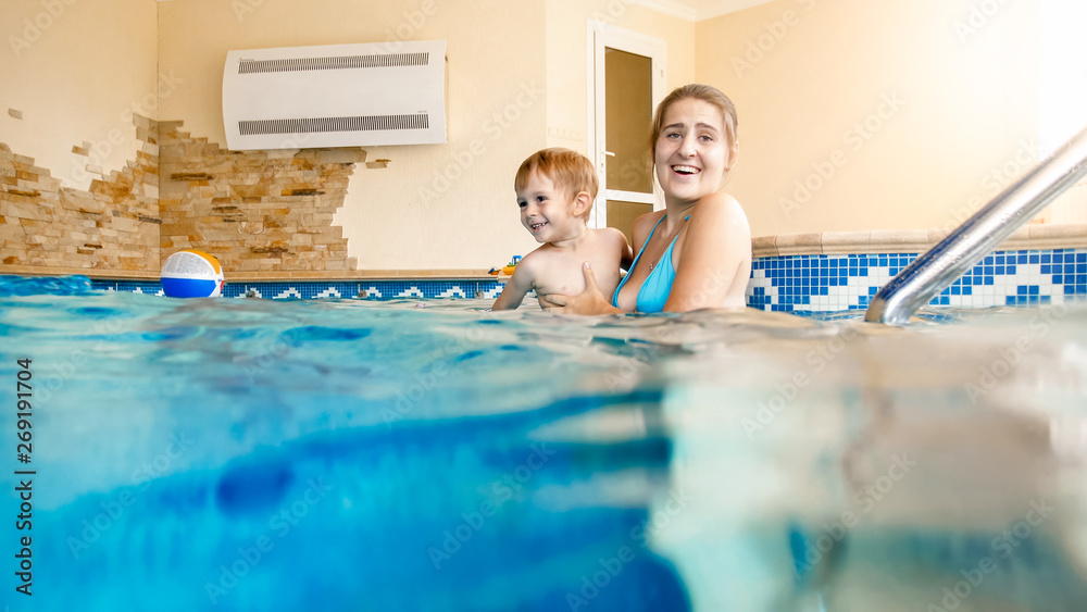 Portrait of happy cheerful young mothet with 3 years old toddler boy playing in the pool at house. Child learning swimming with parent. Family having fun at summer