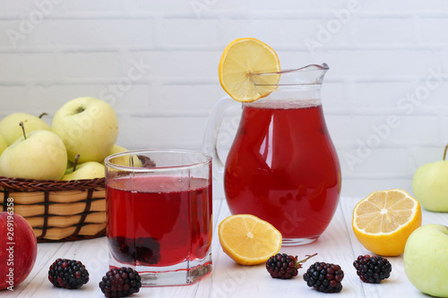 Compote of berries and apples in a jug and in a glass on a table on a white background. In the photo there are apples, blackberries and lemon