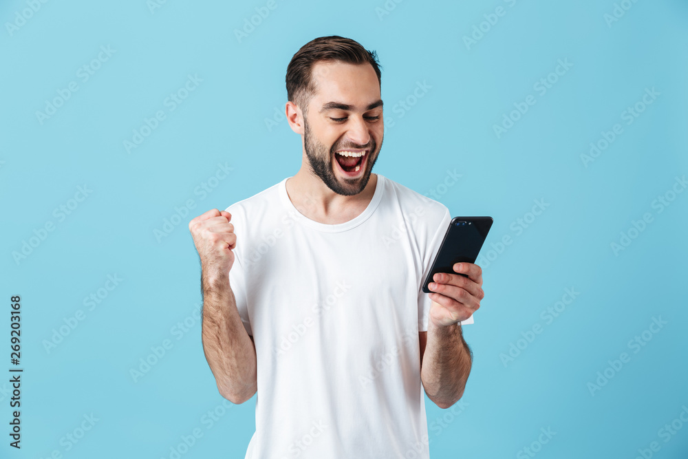 Photo of attractive brunette man wearing basic t-shirt laughing and holding smartphone - obrazy, fototapety, plakaty 