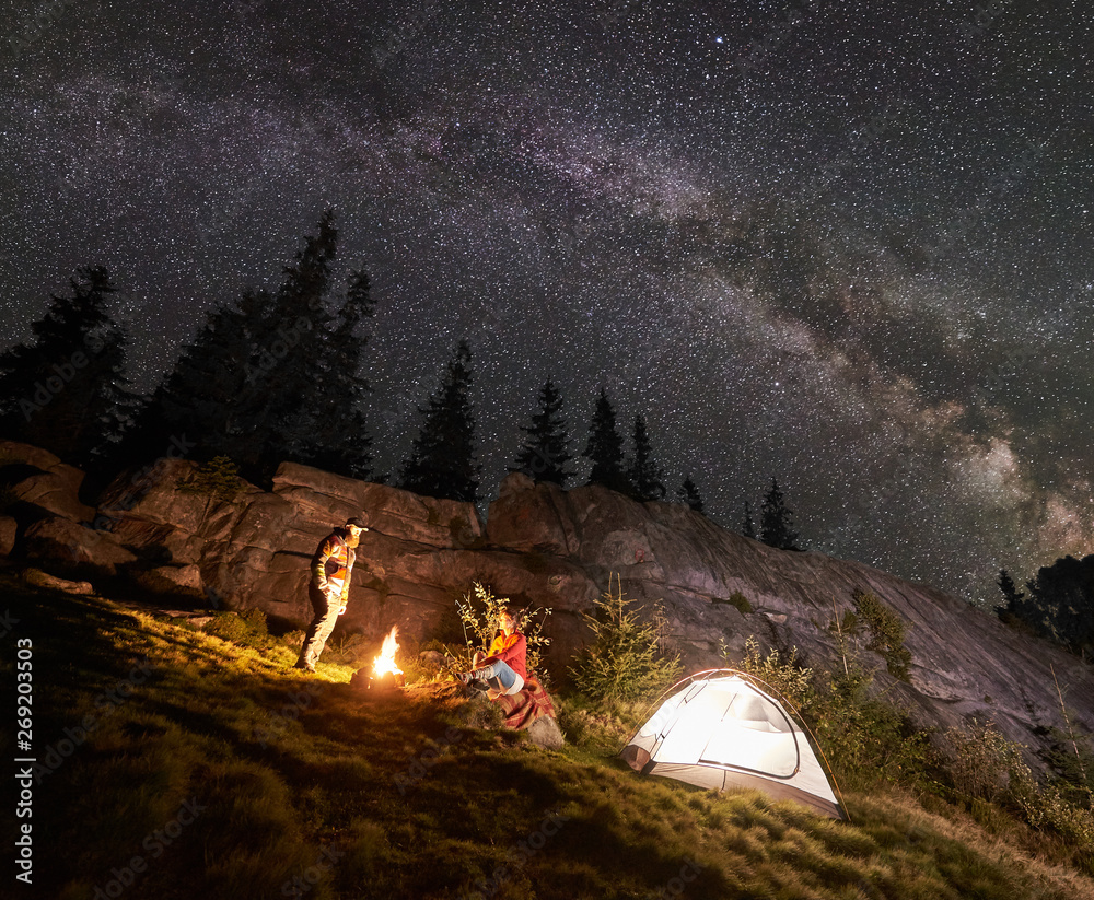 Night summer camping in mountains. Young couple hikers having a rest together beside bonfire and glowing tourist tent under starry sky full of stars and Milky way. On background big boulder and trees. - obrazy, fototapety, plakaty 