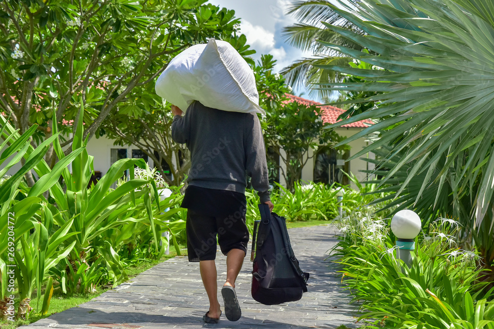 A man in a gray sweater and black shorts carries a white bag on his shoulder and holds a backpack in his hand. He walks along a stone path among palm trees and green plants around the hotel