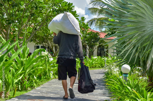 Obraz na płótnie A man in a gray sweater and black shorts carries a white bag on his shoulder and holds a backpack in his hand. He walks along a stone path among palm trees and green plants around the hotel w salonie