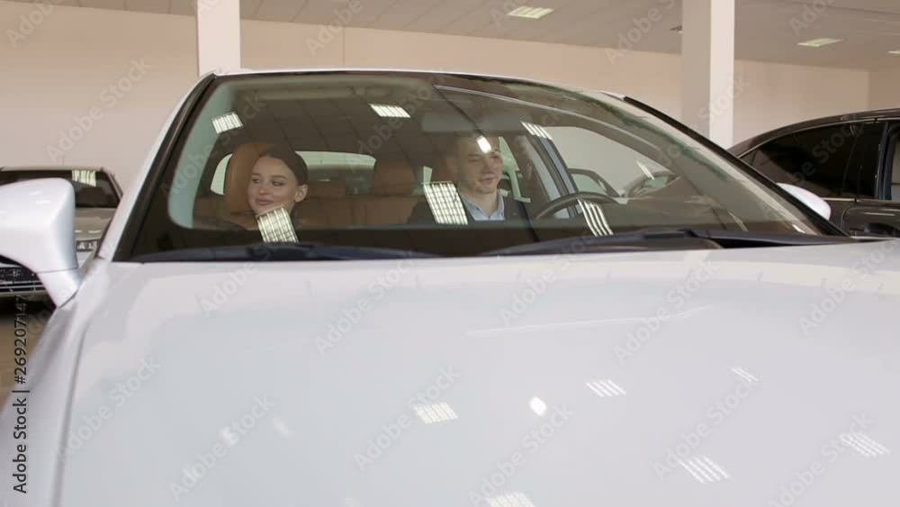Young happy couple chooses a new car in the showroom, they sit in the car and consider it. Young couple watching a new white car in the showroom of the dealership. Young happy couple chooses a new car in the showroom, they sit in the car and consider it. Young couple watching a new white car in the showroom of the dealership.
