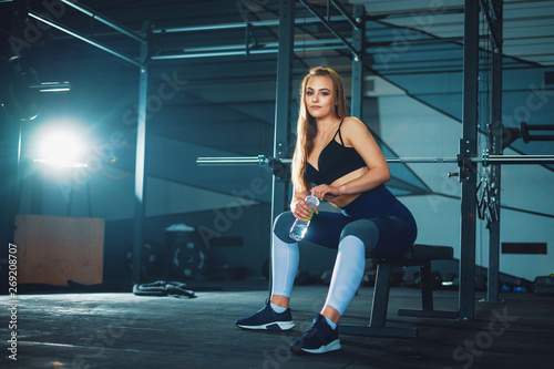 Young attracrive sport woman sitting with water bottle on crossfit gym background