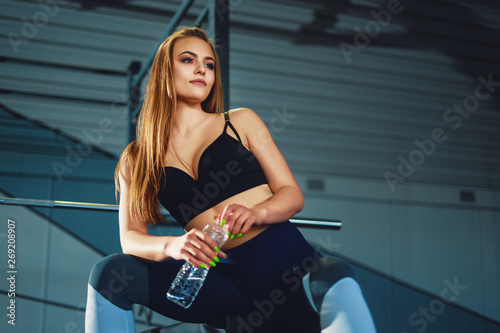 Young attracrive sport woman sitting with water bottle on crossfit gym background