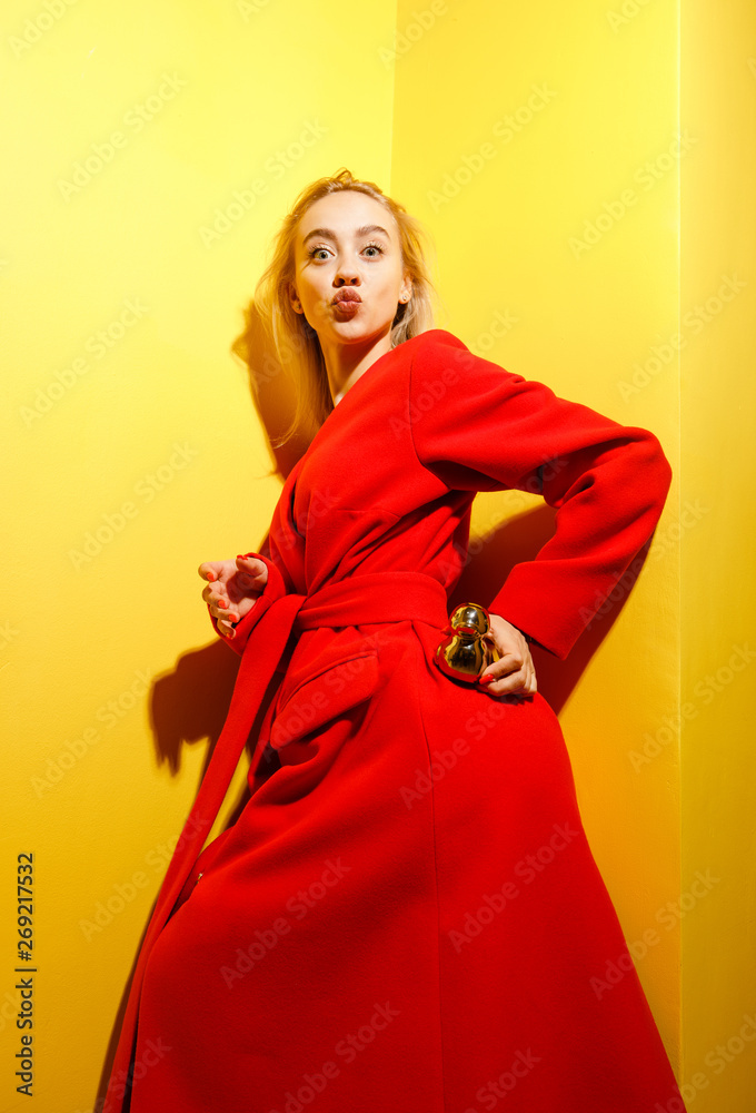 Fototapeta premium Fashion young girl blogger dressed in stylish red coat poses with the gold little duck figurine in her hands on the background of yellow walls in the show room