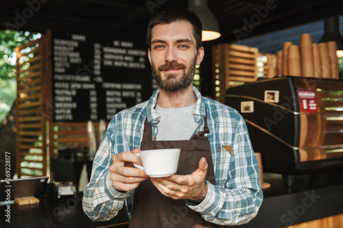Portrait of smiling barista man holding cup of coffee while working in street cafe or coffeehouse outdoor