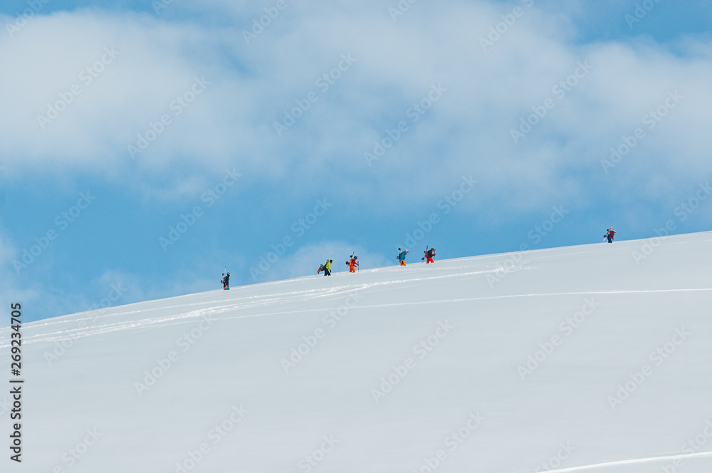 A group of snowboarders climbing the mountain ridge with their boards on the back and with clouds as a beautiful background
