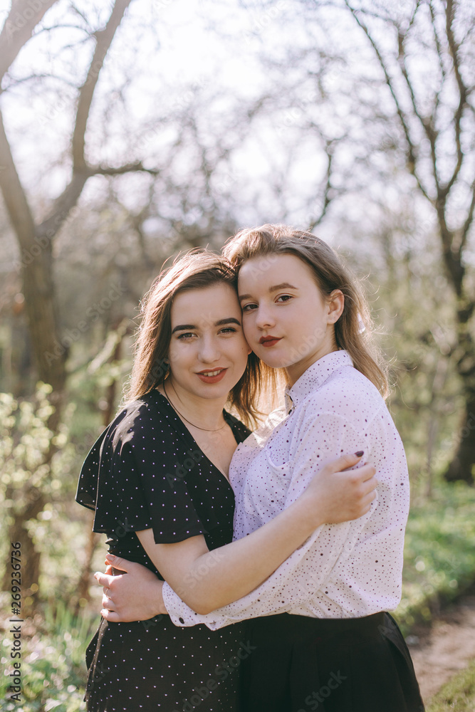 Fototapeta premium Portrait of two beautiful young sisters in the green spring garden, hugging in the field in the sunshine. Having fun together, positive emotions, bright colors. Copy space. Happy girlfriends at sunset
