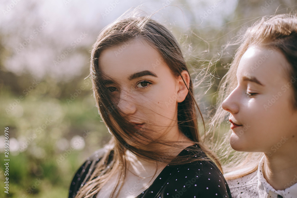 Portrait of two beautiful young sisters in the green spring garden, hugging in the field in the sunshine. Having fun together, positive emotions, bright colors. Copy space. Happy girlfriends at sunset