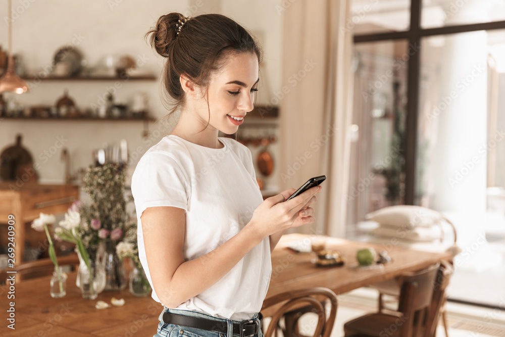Image of optimistic nice woman typing on cellphone and smiling in cozy kitchen Image of optimistic nice woman typing on cellphone and smiling in cozy kitchen