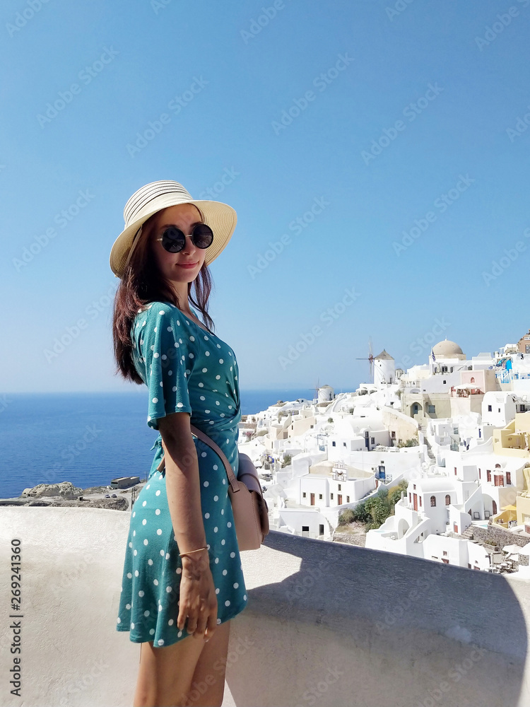 Beautiful young woman in green dotted dress walking in Oia, Santorini streets. White houses, blue sea