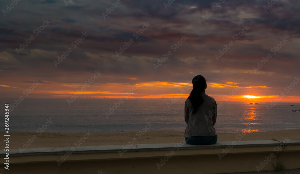 Unidentifiable woman in silhouette, sitting alone watching sun set over ocean at beach. Dramatic sky with vivid colors.