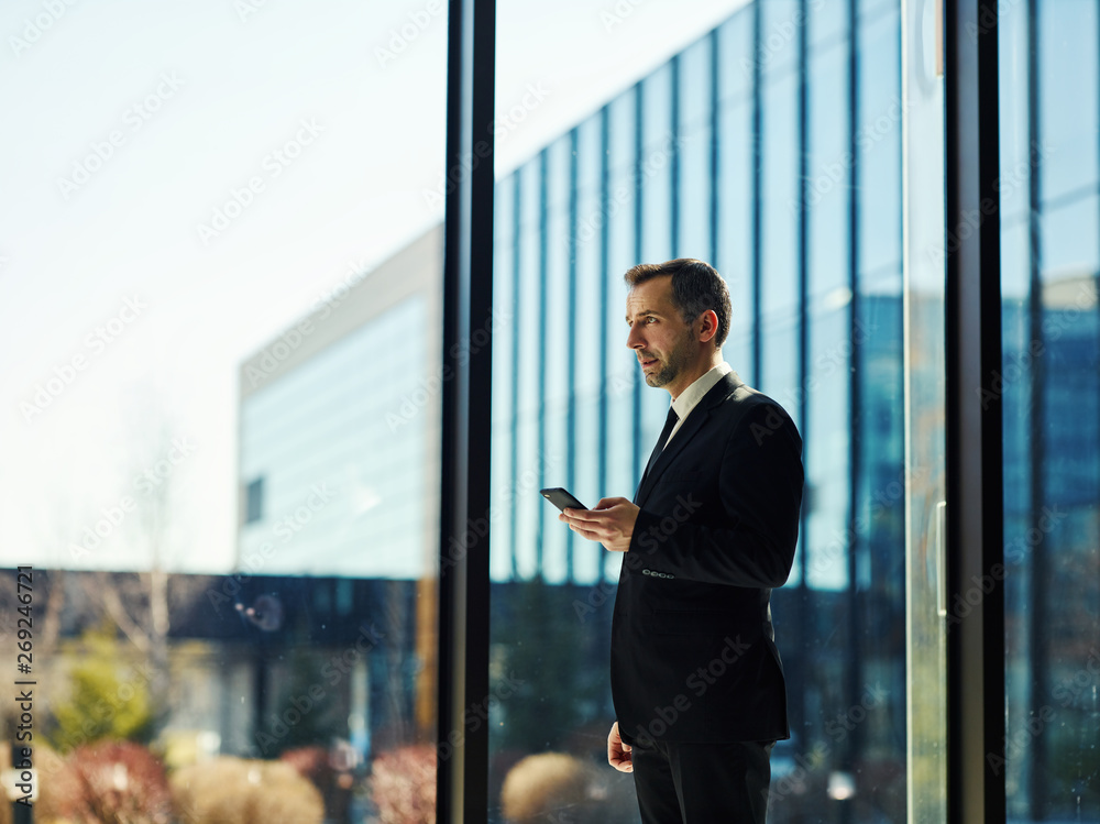 Side view of confident manager in black suit standing by floor length window, holding mobile phone and looking away thoughtfully Side view of confident manager in black suit standing by floor length window, holding mobile phone and looking away thoughtfully