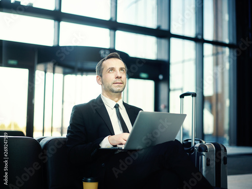 Businessman in suit sitting on leather sofa in airport lounge and looking at departure board, laptop on his lap and trolley suitcase placed nearby