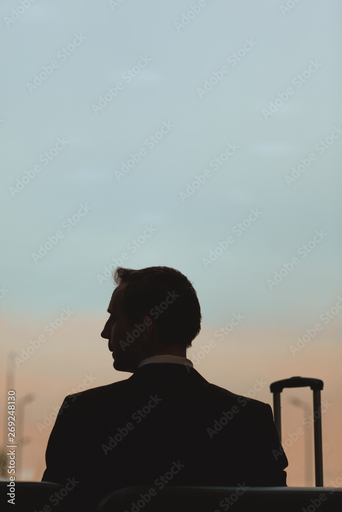 Silhouette of businessman sitting on bench in airport with suitcase nearby waiting for flight and thinking of future Silhouette of businessman sitting on bench in airport with suitcase nearby waiting for flight and thinking of future