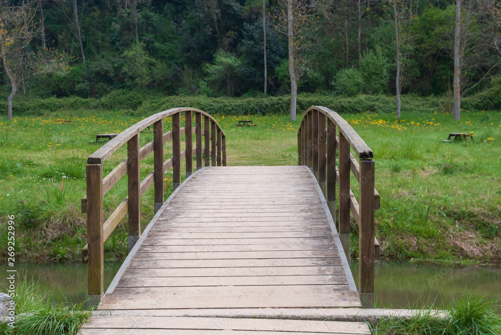 Wooden bridge over a small river with vegetation and a picnic area in the background in Cobreces, Cantabria, Spain, Europe