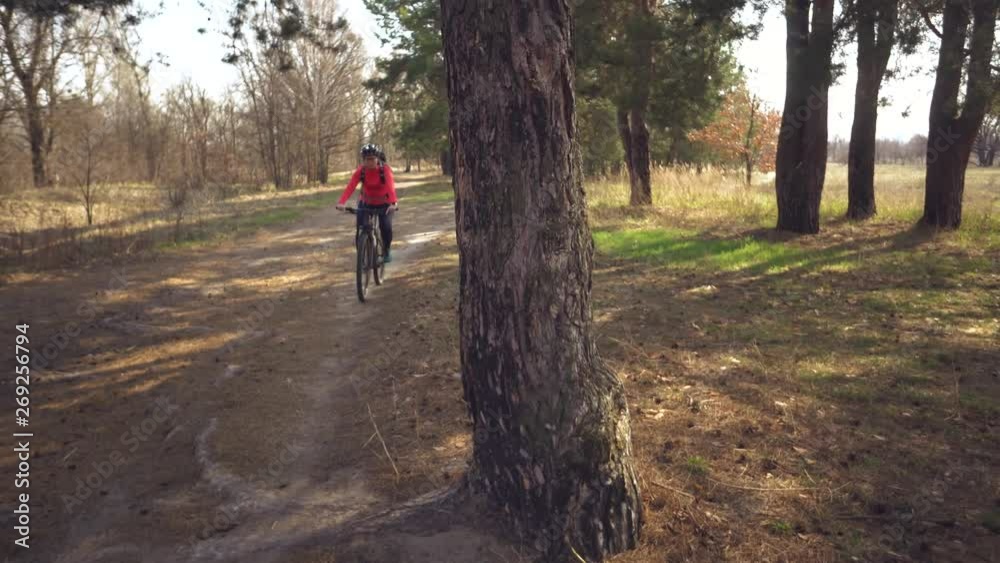 Caucasian sport cyclist woman sport off-road bike park near tree. A female athlete in sportinvnoy clothing and a helmet and a black backpack break stop training on mountain bike Caucasian sport cyclist woman sport off-road bike park near tree. A female athlete in sportinvnoy clothing and a helmet and a black backpack break stop training on mountain bike