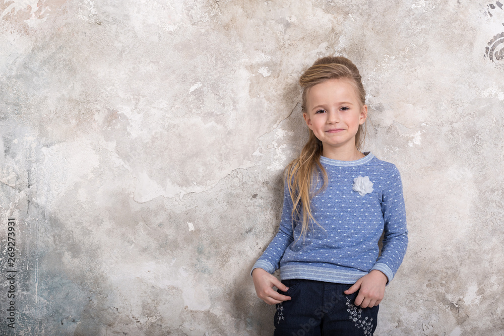 Portrait of a little attractive smiling girl in a blue sweater and pants with hair folded into her hair against a grunge wall background. Portrait of a little attractive smiling girl in a blue sweater and pants with hair folded into her hair against a grunge wall background.