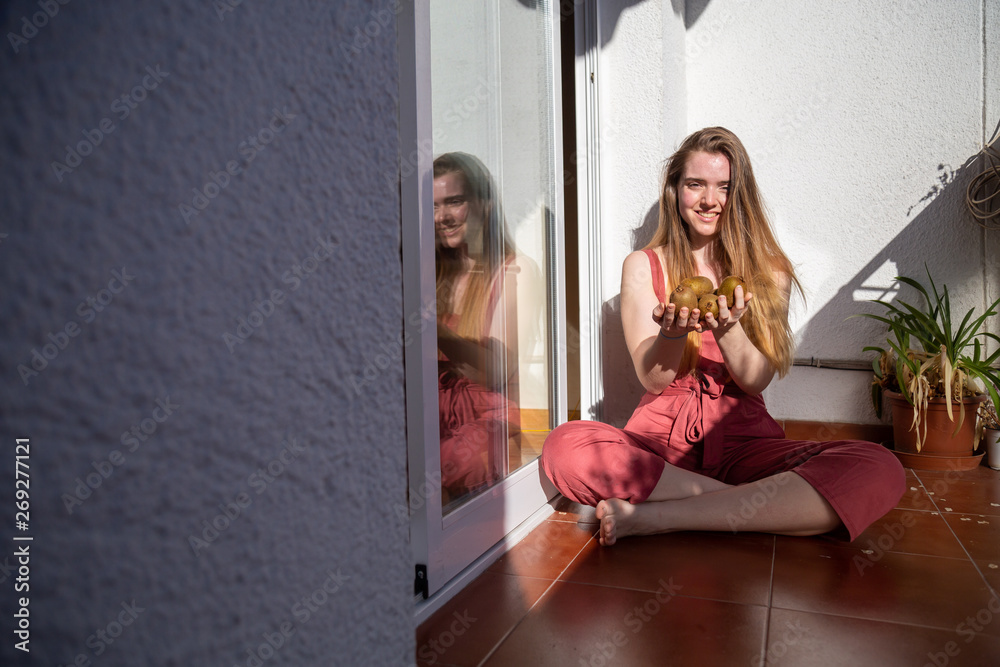 Young charming female in casual summer dress sitting on balcony and offering tropical fresh juicy kiwis Young charming female in casual summer dress sitting on balcony and offering tropical fresh juicy kiwis