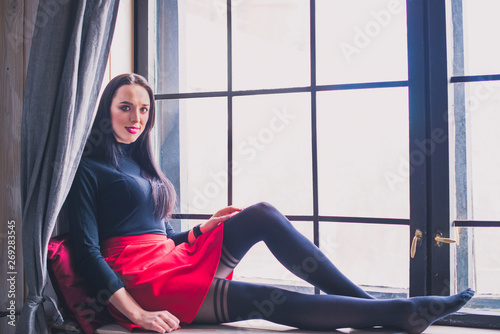 A beautiful, young brunette model in a black turtleneck and a red skirt is sitting on the windowsill.