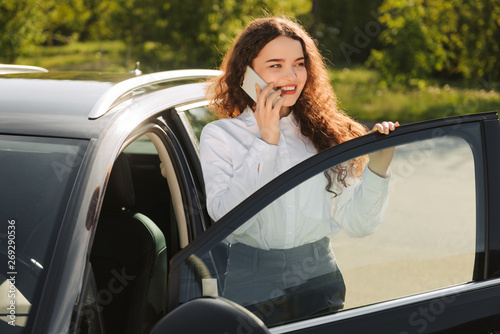 Business woman talking on the phone in the parking lot. Stylish young woman standing near the car © JJ Studio