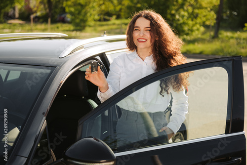 Outdoor shot of satisfied beautiful female owner of car, likes and rejoices her purchase, holds keys , has happy expression, stands near new automobile, going to drive © JJ Studio
