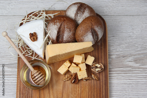 Top view of hard and brie cheeses of in assortment with nuts, honey and buns on a wooden board and light background. The concept of still life. Space for text
