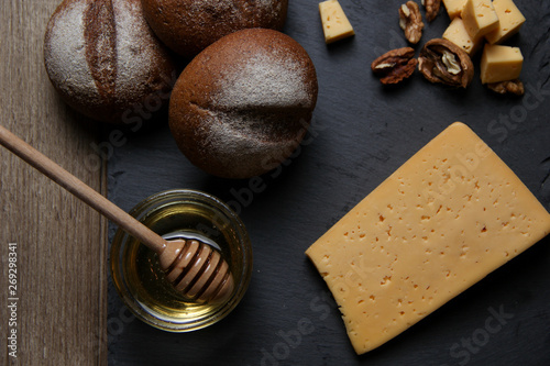 Top view of hard and brie cheeses of in assortment with nuts, honey, buns on a black stone board and wooden background. The concept of still life. Space for text