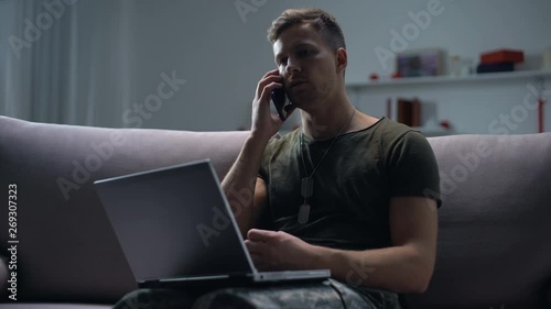 Male military spy sitting with laptop on sofa and talking smartphone at night photo