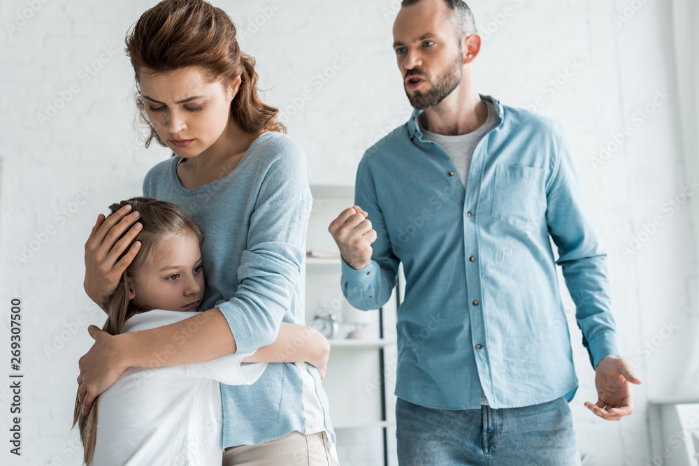 Fototapeta premium upset mother hugging daughter while angry man showing fist at home