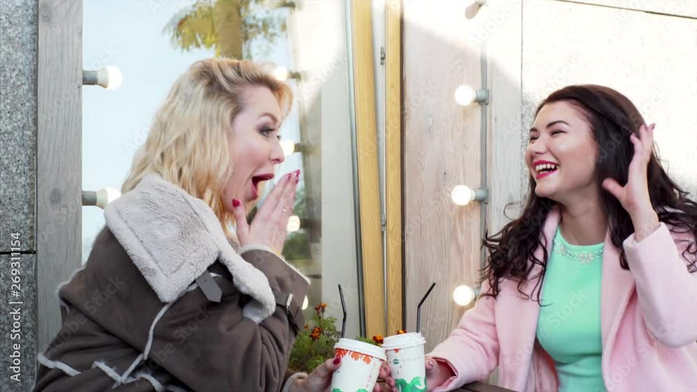 Two attractive women burst out laughing sitting at street cafe with coffee and having fun Two attractive women burst out laughing sitting at street cafe with coffee and having fun