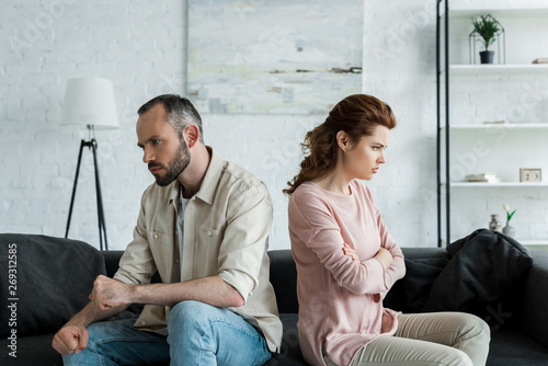 upset woman sitting near angry husband with crossed arms at home