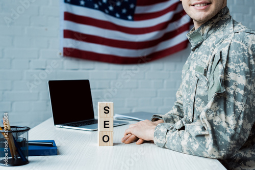 cropped view of happy soldier near wooden cubes with seo lettering and laptop with blank screen
