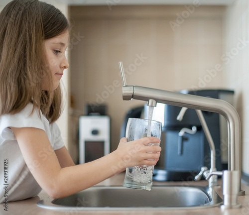 Portrait of a little caucasian girl gaining a glass of tap clean water. Kitchen faucet. Cute curly kid pouring fresh water from filter tap. Indoors. Healthy life concept