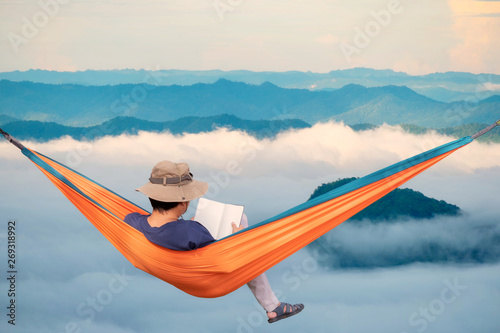 View of a man in hammock at sunrise with dense fog nature background. Young man reading a book lying in a hammock.